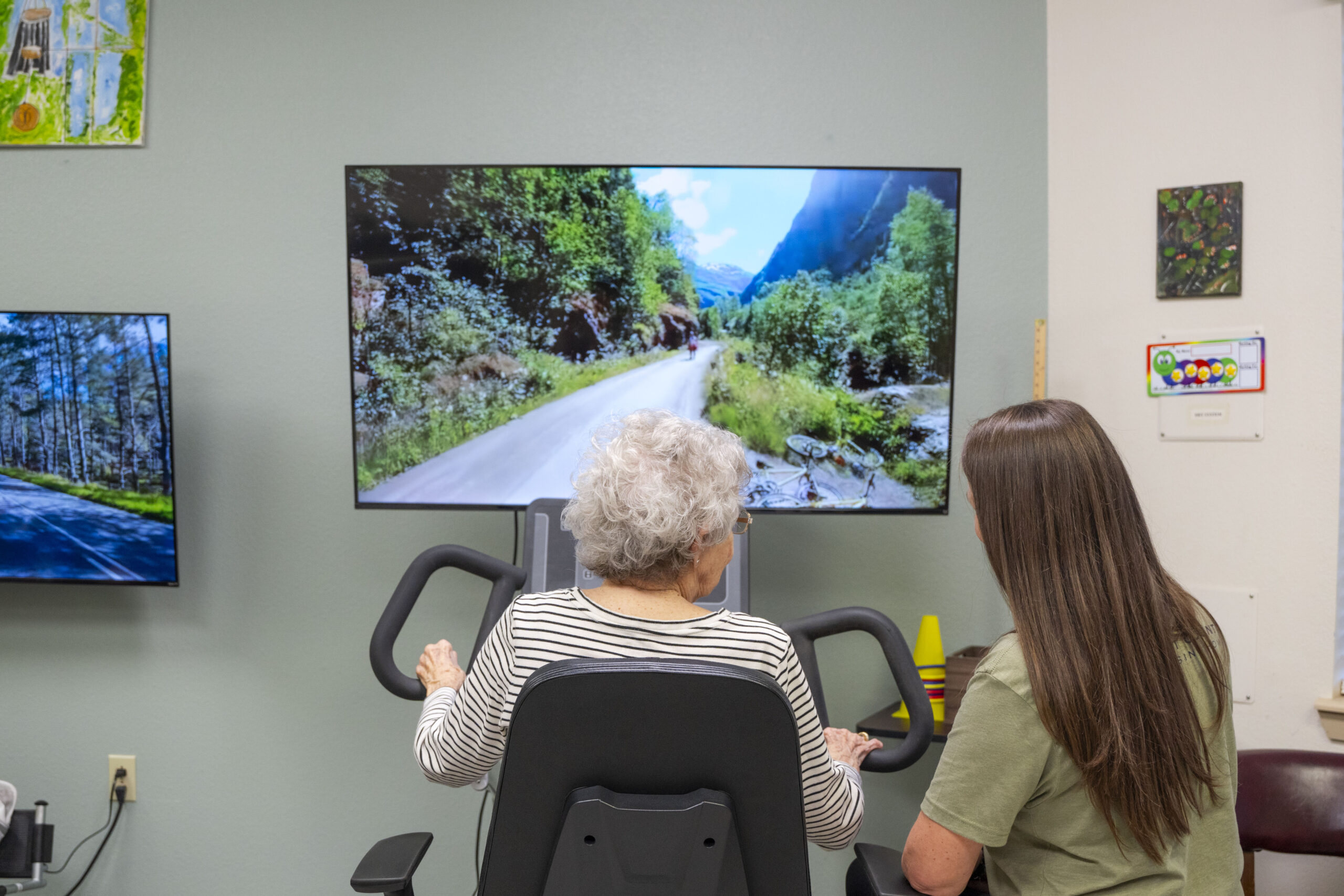 Resident smiling while doing leg exercises with supportive staff at Kent County Nursing Home in Jayton, TX. A TV screen in front of the machine depicts a biking trail that the machine is mimicking